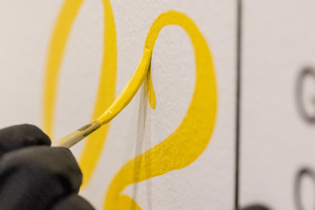 A hand holds a paintbrush tracing bright yellow shapes on a white surface.