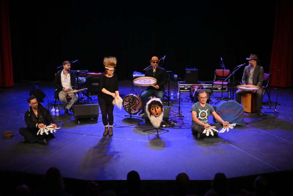 Six people on a spotlighted stage; three play traditional Inuit percussion instruments, and three engage in dance and movement.
