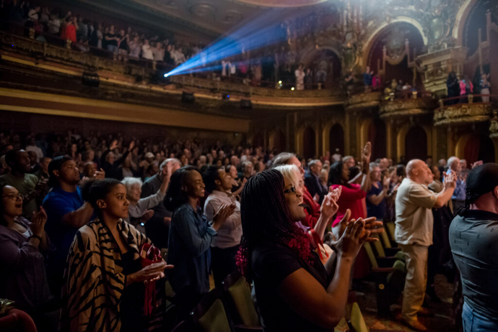 Inside of a beautifully adorned theater, a full audience stands and applauds.