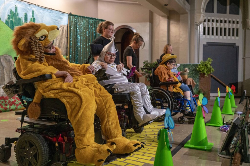 Six people on stage, three wearing costumes of a lion, tin man, and scarecrow.