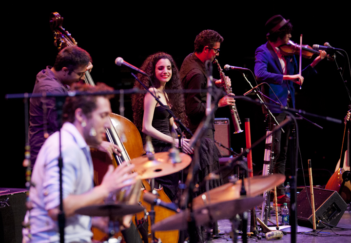 A vibrant ensemble performs on stage under dramatic lighting. A woman in a black dress smiles at the audience while fellow band members play upright bass, drums, clarinet, and violin.