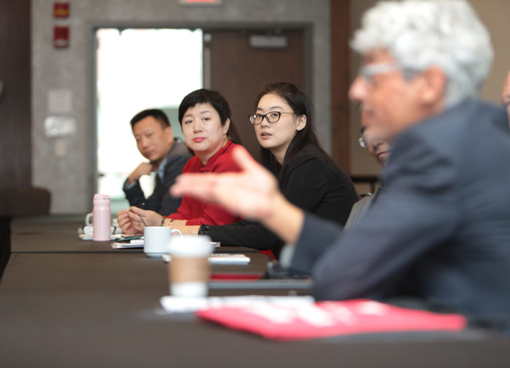 The focus is on a group of people sitting at a long table in a session room, looking at the camera, and close up and out of focus there is a person gesturing and looking forward.