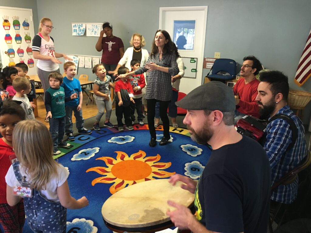 Musicians perform for a lively group of preschool-aged children, who dance and sing along while standing on a colorful rug decorated with numbers and a sun.