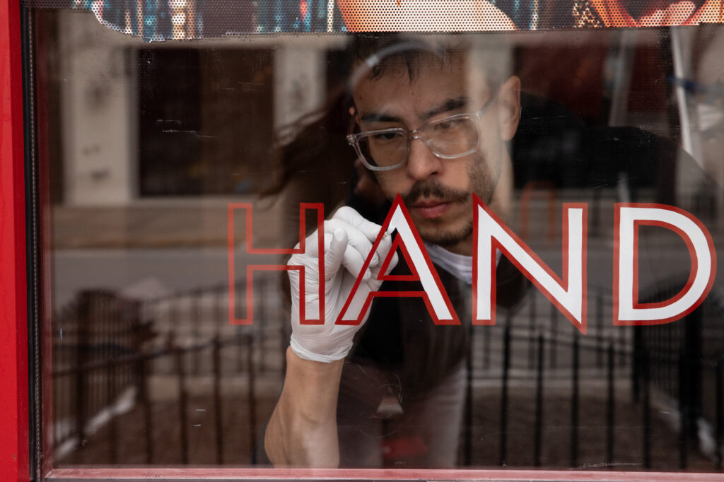 A person with medium-light skin tone wearing clear glasses and a white glove paints a letter "A" on a window.