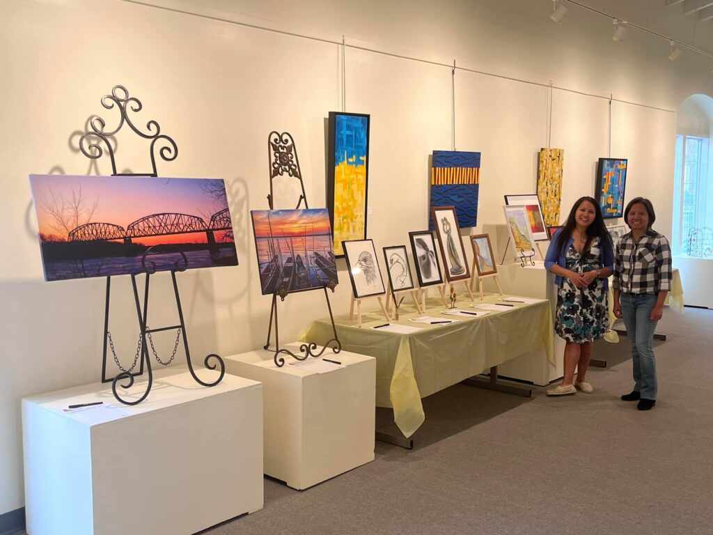 Two women stand in front of a display of art in an indoor gallery.
