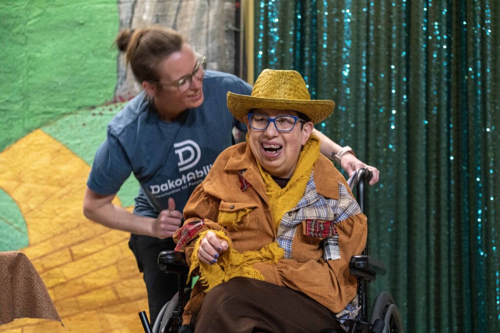 A light-skinned man wearing a scarecrow costume smiles on stage while a light-skinned woman wearing blue assists from behind.
