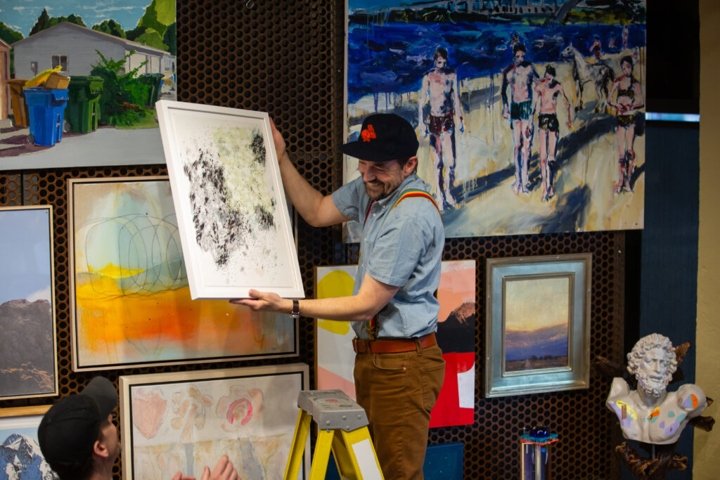 A person standing o a ladder next to a wall filled with artworks. They are handing off an artwork from the wall to a person standing below them.