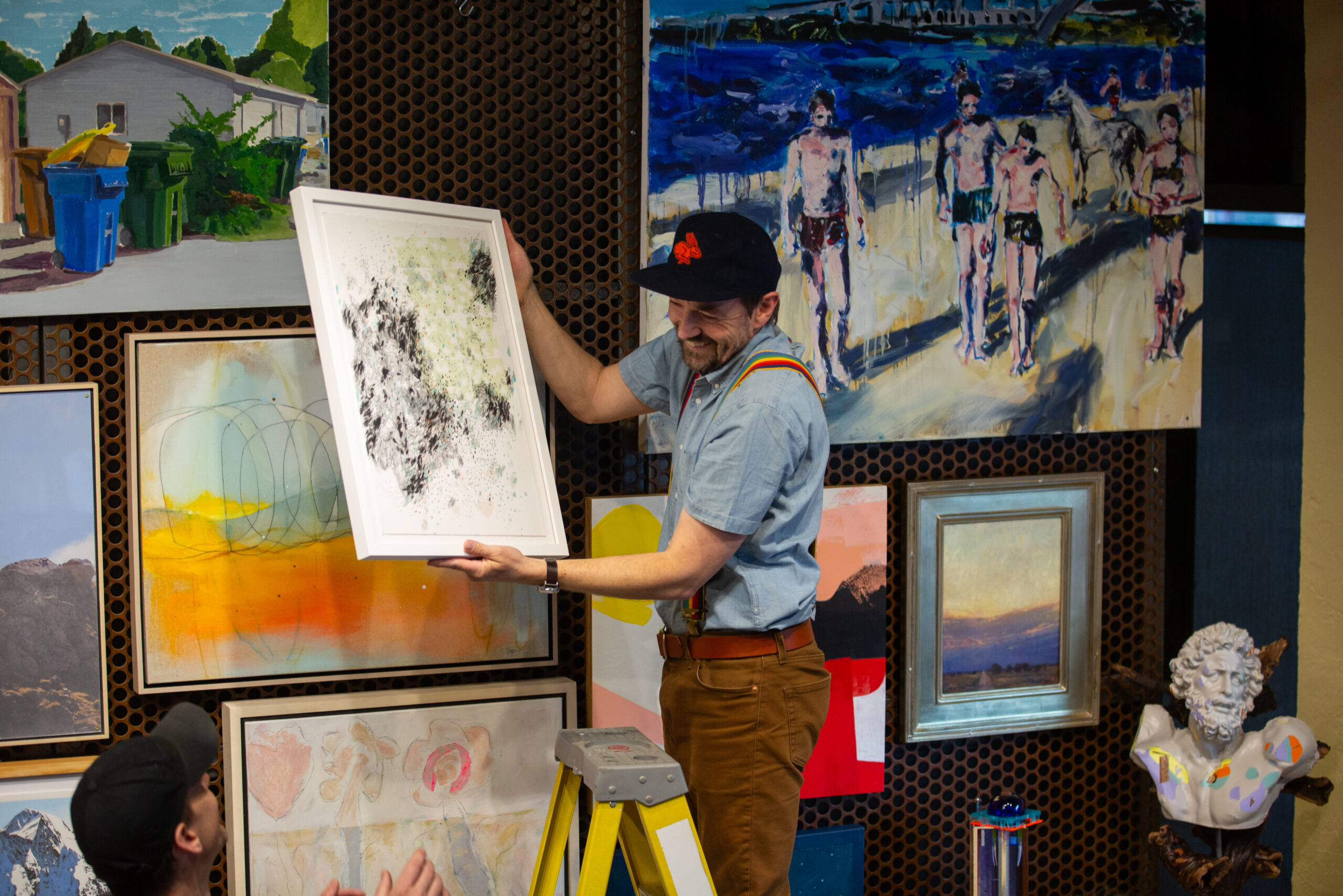 A person standing o a ladder next to a wall filled with artworks. They are handing off an artwork from the wall to a person standing below them.