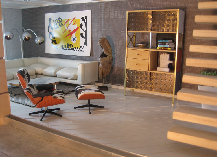 The inside of a miniature house with mini living room furniture including an L-shaped couch, bookshelf, and artworks