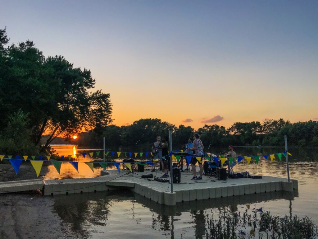 A small band performs on a floating dock at sunset, with colorful triangular pennant flags strung across the dock. The orange sun sets over a calm river behind them.