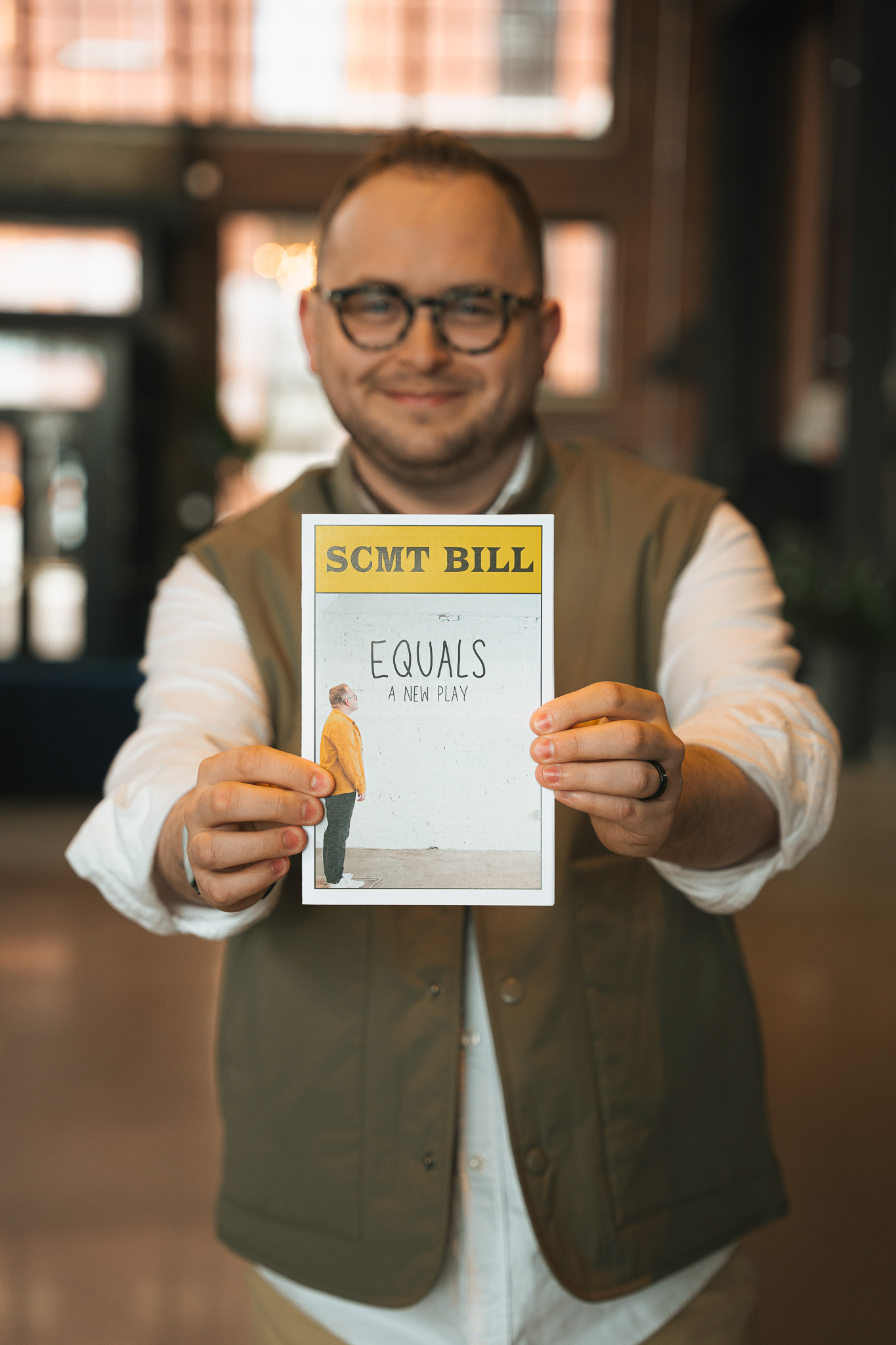 A man with glasses and a khaki vest holds a playbill up to the camera. On it is the title "Equals" and a singular figure looking up at the word.