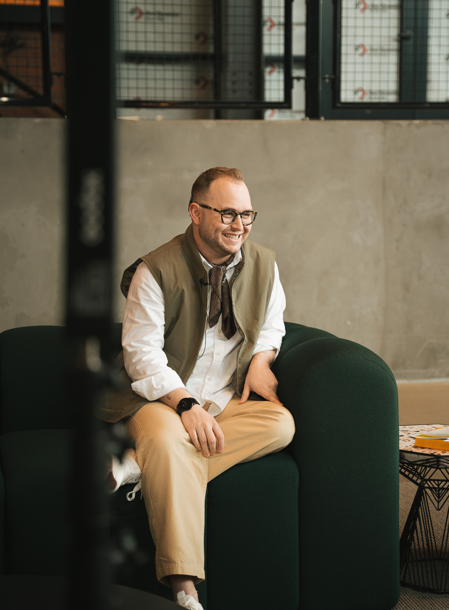 A smiling person wearing glasses, khaki pants, a khaki vest, and an ascot tie sits in a large green chair.