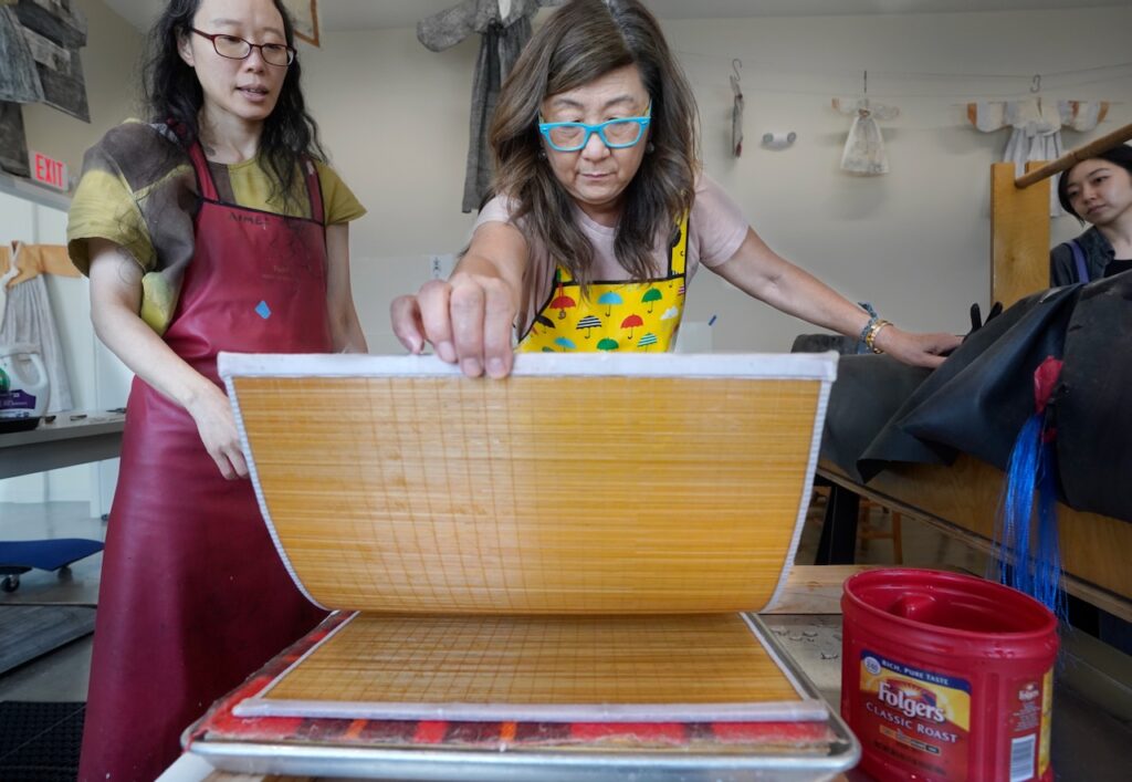 A person with long black hair in a red apron standing next to a person with long brown hair in a yellow patterned apron as the center person pulls a bamboo screen away from a table next to a vat, in a papermaking studio. Watching from the vat is another person with black hair.