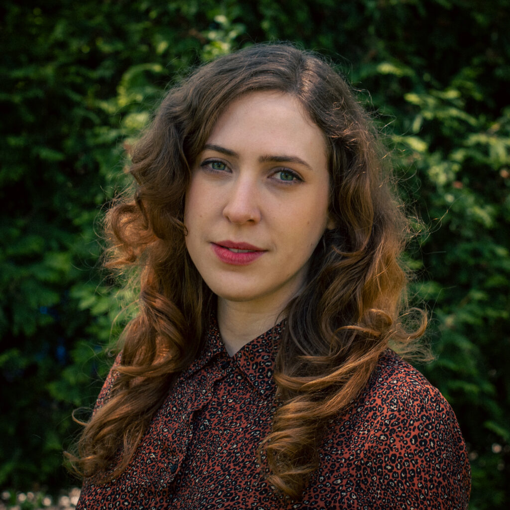 A woman with long, wavy brown hair stands outdoors in front of lush green foliage. She wears a patterned button-up blouse and looks directly at the camera with a calm expression.
