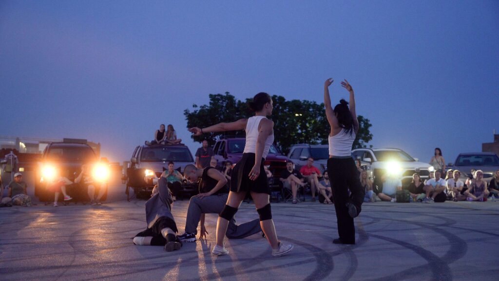 Four dancers performing in a parking lot as the people watch from their parked cars with headlights turned on.