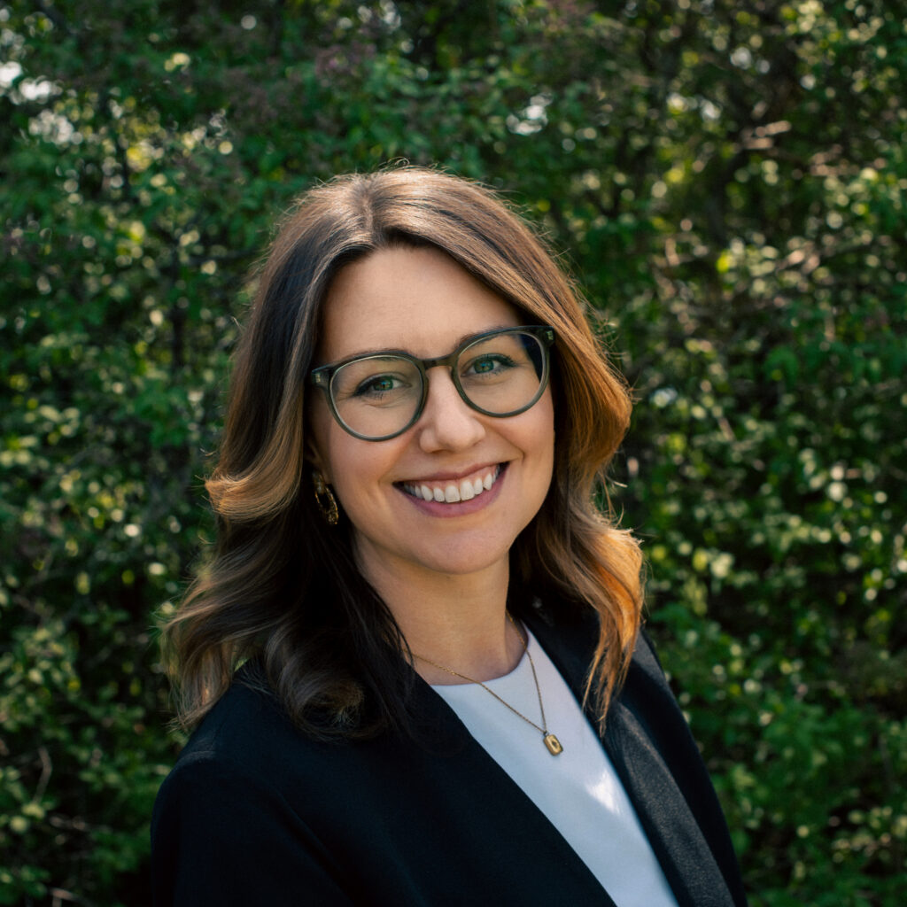 A woman with shoulder-length, wavy brown hair smiles at the camera. She is wearing glasses, a black blazer over a light-colored top, and a gold pendant necklace. The background is filled with greenery and soft sunlight filtering through the trees.