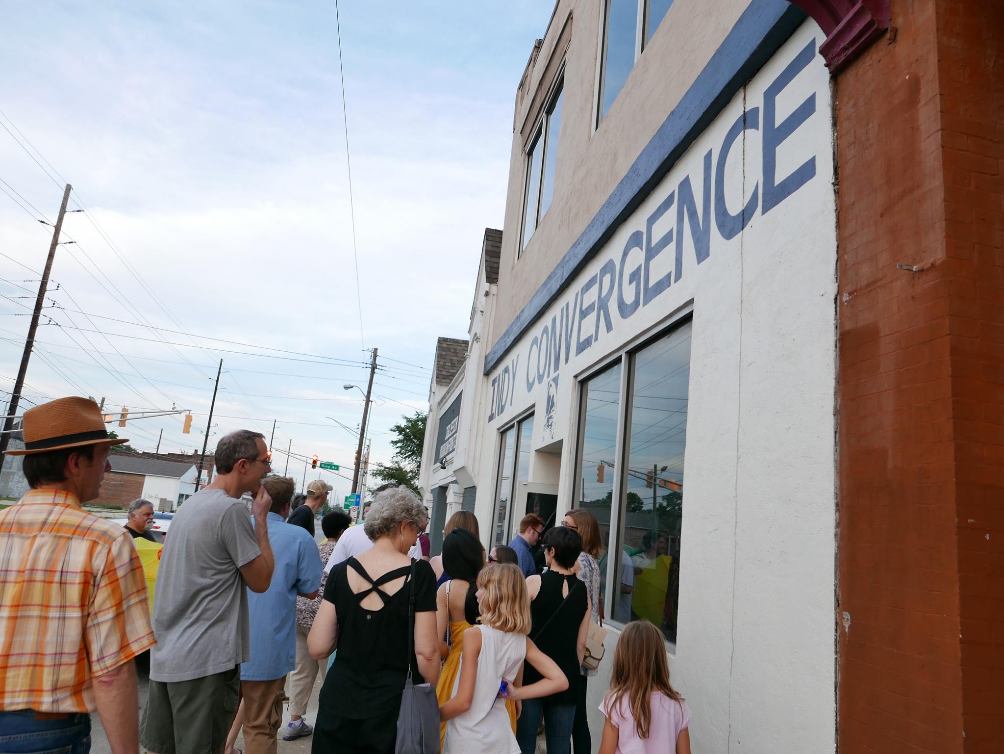 A crowd of people stand in line outside the Indy Convergence building on a sunny day, waiting to enter. The white-painted building has large windows and a sign with blue block letters.