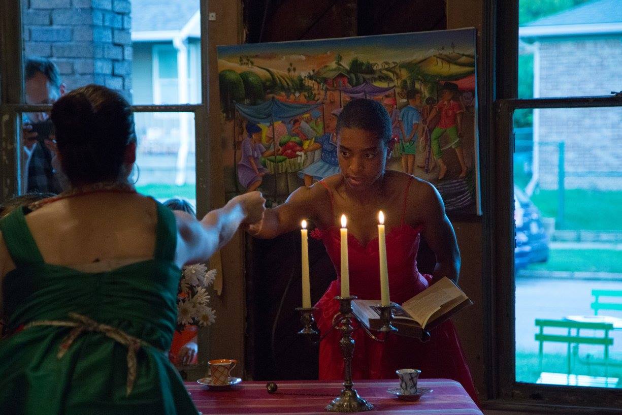 A woman in a red dress holds a book and reaches out toward another person across a table with lit candles, teacups, and a striped tablecloth. A colorful market scene painting hangs behind her.