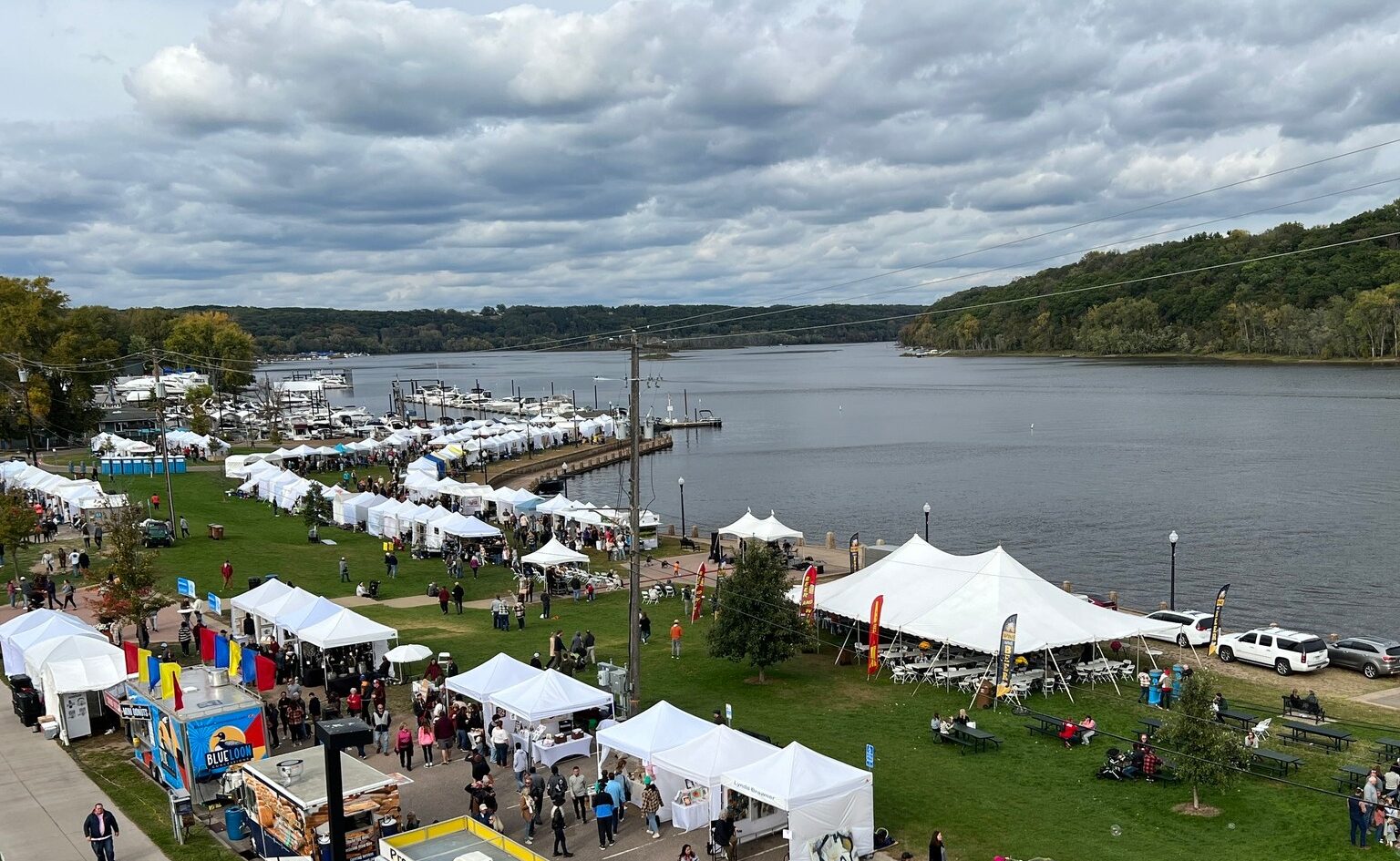 Tents and food trucks are set up along a river on a partly-cloudy day.