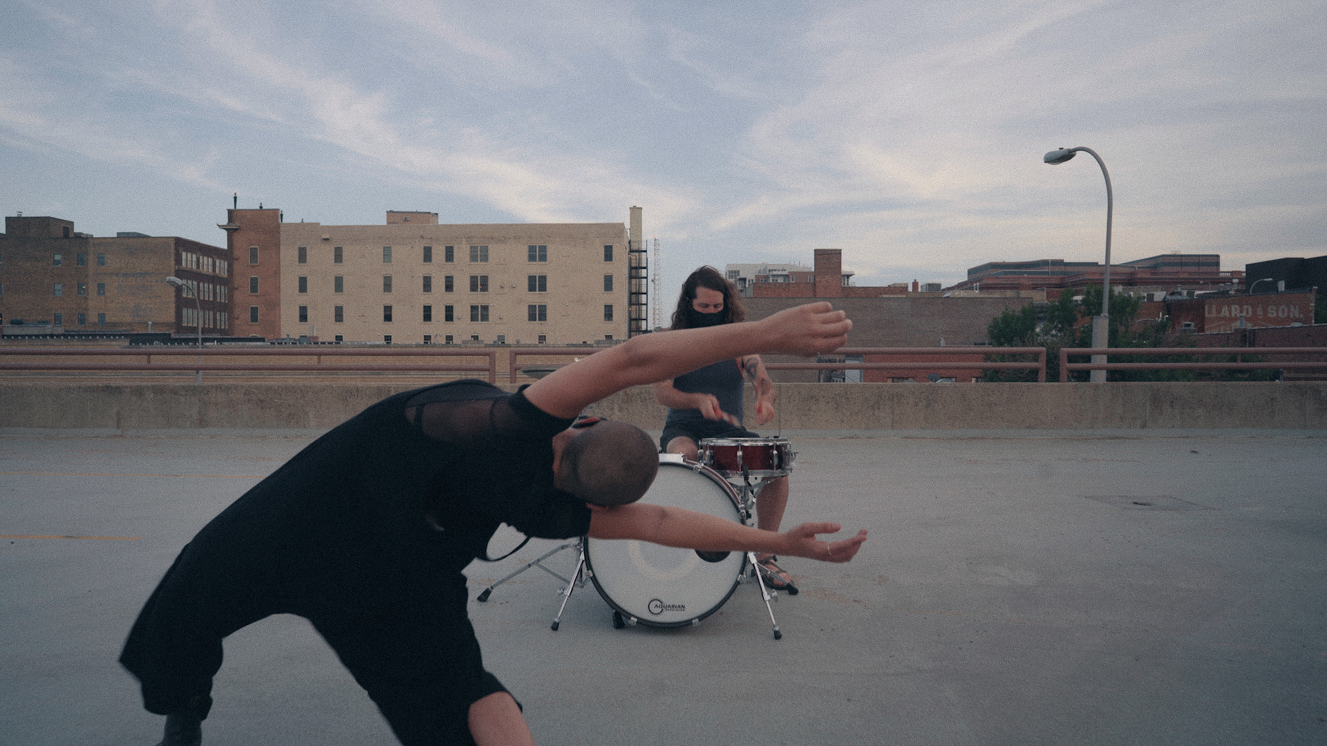 A drummer and a dancer performing in an empty parking lot. Behind them is skyline with buildings.