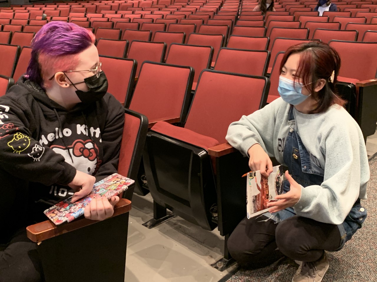 Two people in masks sit in an auditorium filled with red theater seats