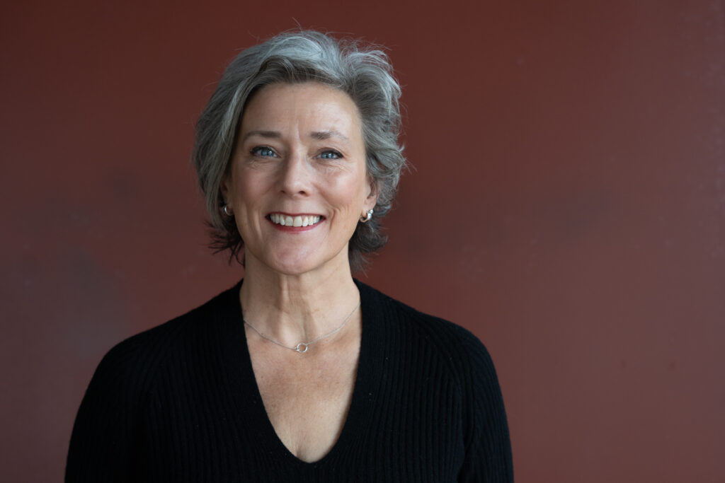 A smiling person with light tone skin and gray hair, wearing a black blouse in front of a brown background.