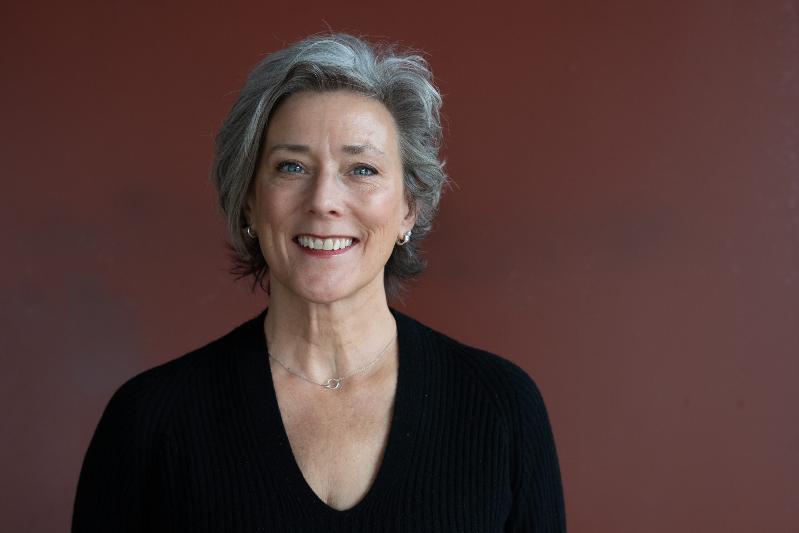 A smiling person with light tone skin and gray hair, wearing a black blouse in front of a brown background.