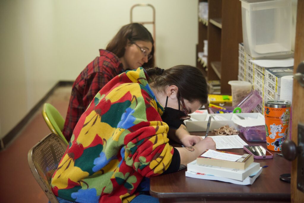Two people writing on pieces of paper while sitting at a desk together.