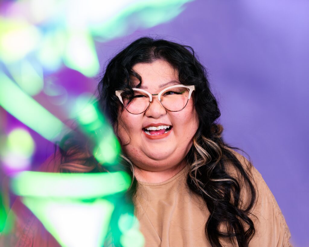 A person of medium light skin tone with long wavy black hair and glasses wearing a tan blouse in front of a purple background