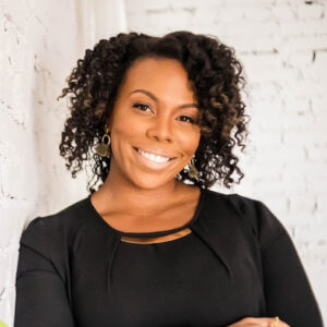 Headshot of a smiling person of medium dark skin tone and short curly black hair with tinges of light brown, wearing gold earrings and a black top, and posing in front of a white brick wall.