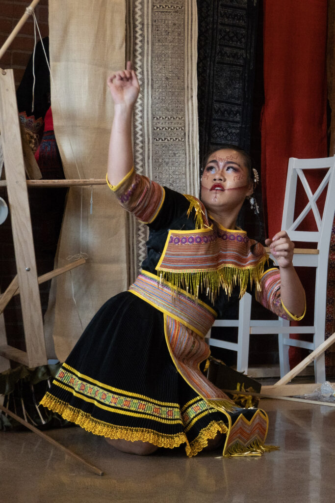 A person in traditional Hmong clothing in the middle of a dance routine in a gallery space.