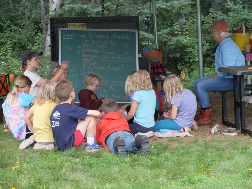A group of children gather in a circle in front of a green chalkboard with a schedule written on it.