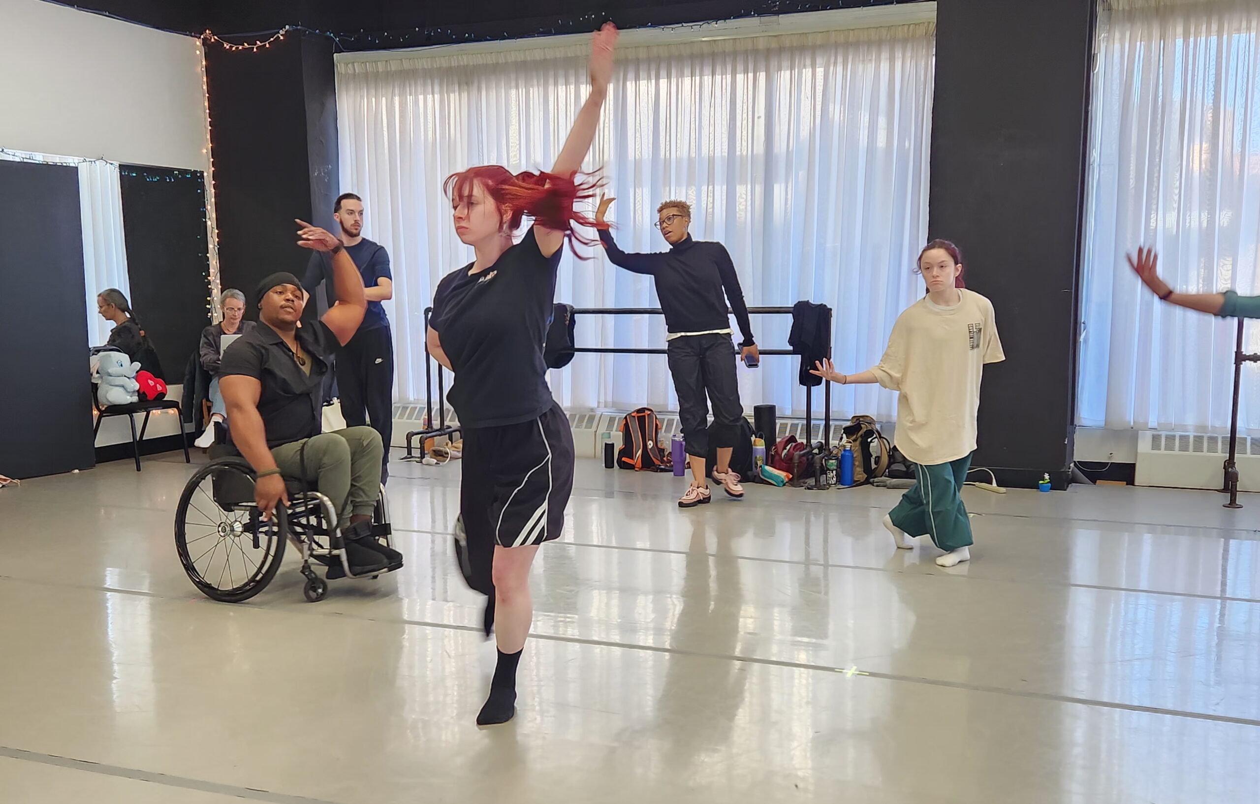 A choreographer, standing with one arm raised in a curved shape, describes a turning arm movement for the dancers to try.