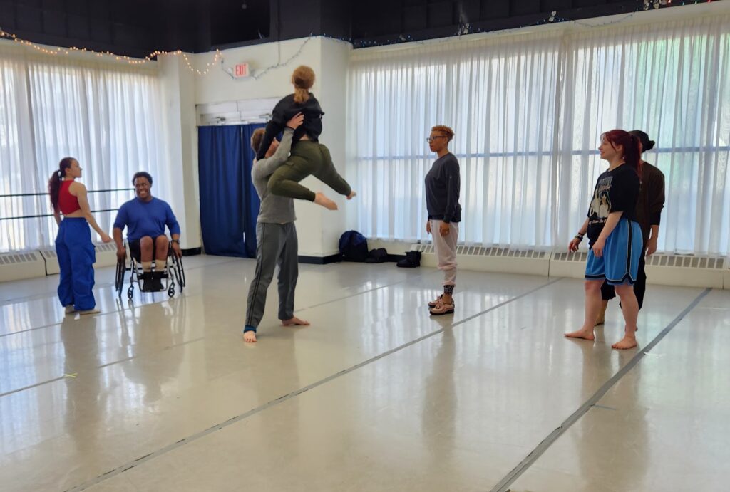 Facing each other, one dancer lifts another, holding a leg extended and the other bent. The choreographer and four dancers look on.