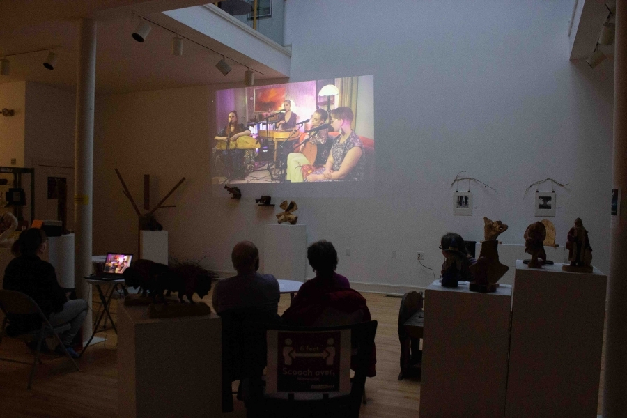 People sit in chairs in a dimly lit gallery, watching a performance being projected on the wall.