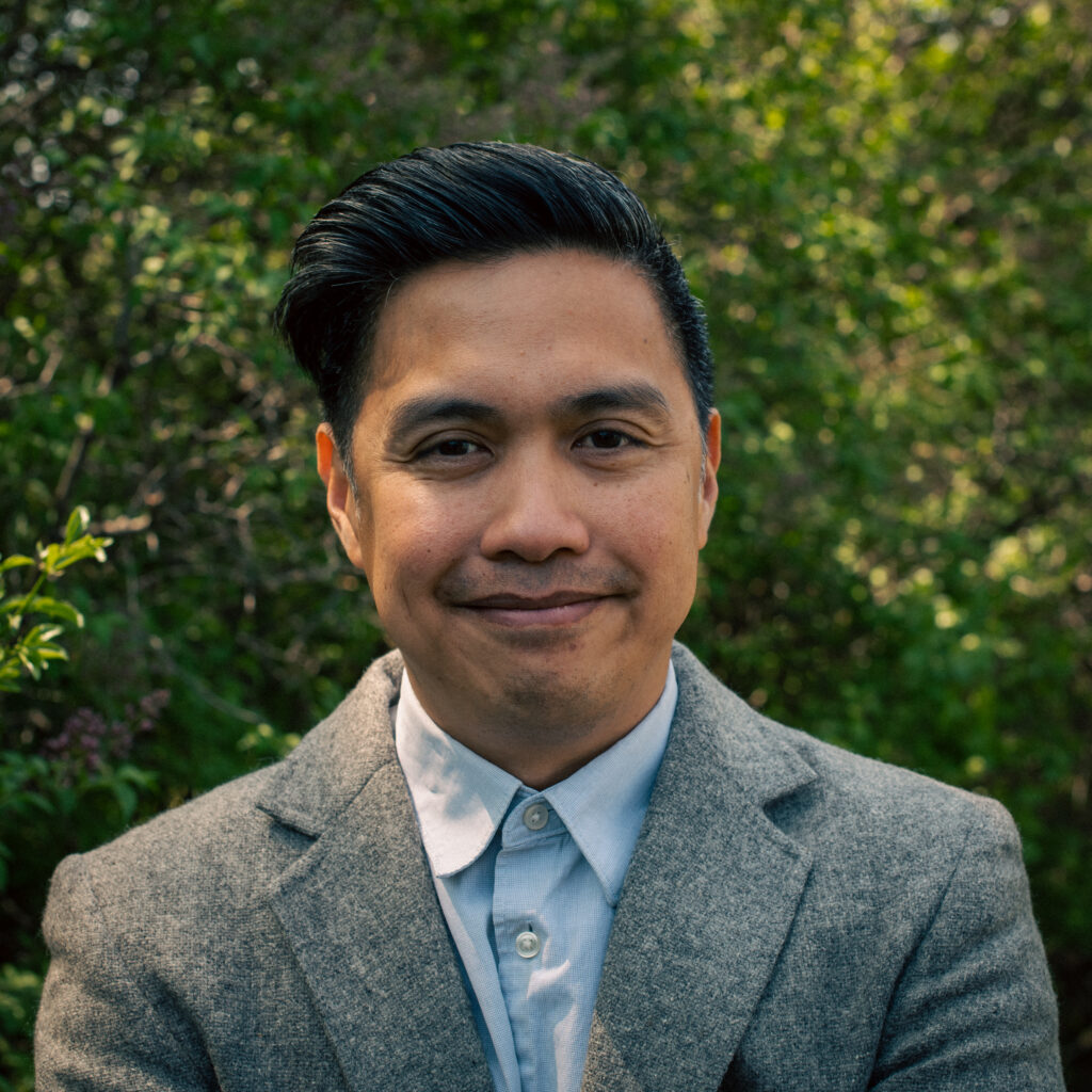 A man with short, neatly styled black hair stands outdoors in front of leafy greenery. He is wearing a gray blazer over a light blue button-up shirt and smiles gently at the camera.
