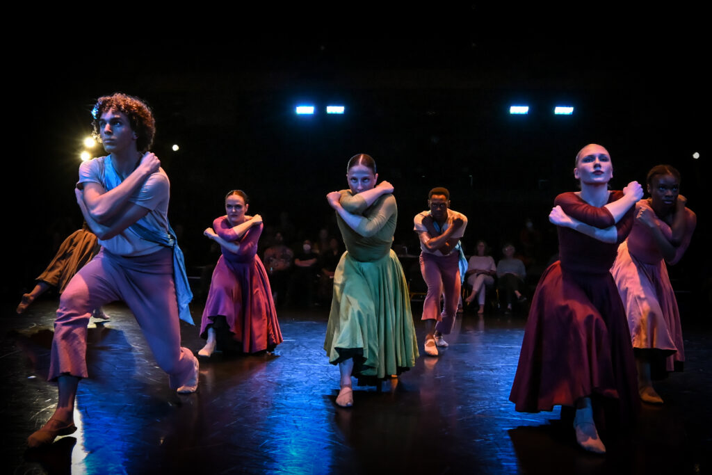 A group of dancers in colorful costumes perform on a dimly lit stage, stepping forward with arms crossed tightly over their chests