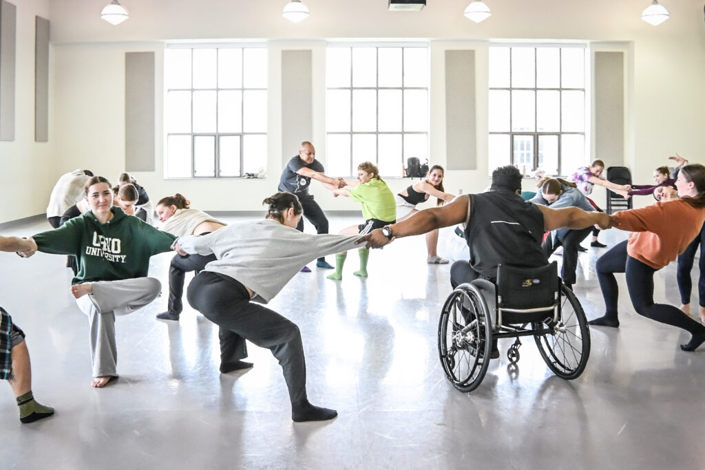 Several dancers fill a brightly lit studio. Most stand in a squat position, and one sits in a wheelchair, as they hold hands with outstretched arms across the studio.