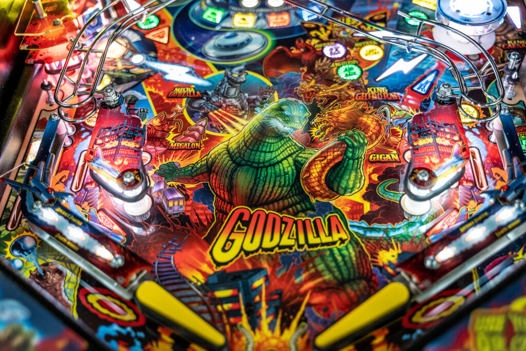 Vivid playfield scene from the Godzilla pinball machine featuring the monster in battle with King Ghidorah, Megalon, and Gigan amid burning buildings and crashing trains.