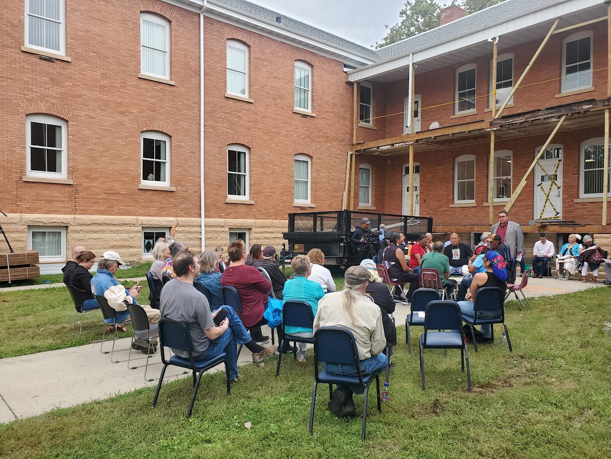 A group gathers outside on chairs listening to a Native drum circle