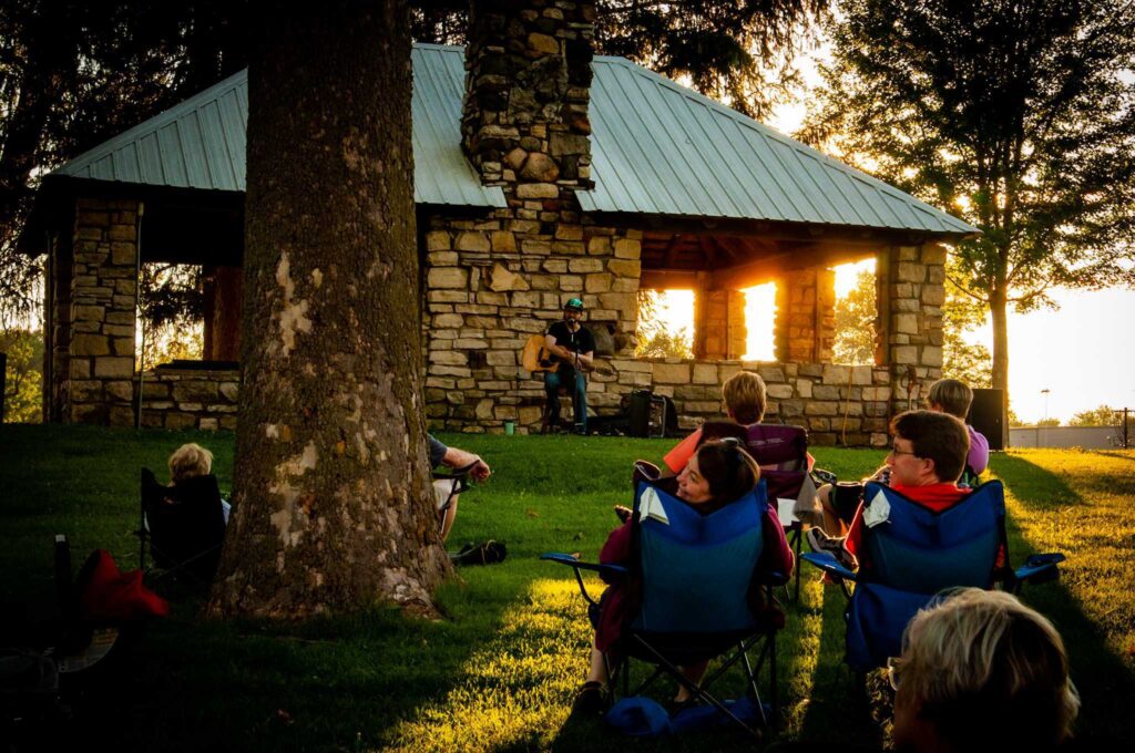 People in folding chairs sit on the grass, facing a person playing guitar and singing into a microphone.