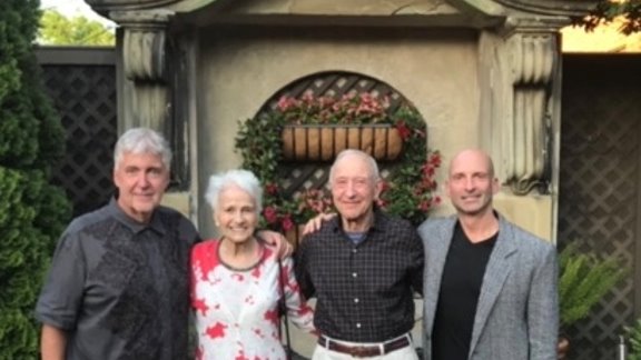 Four adults pose arm-in-arm in front of a decorative outdoor wall with a floral wreath. They smile warmly, standing in a garden or courtyard area with greenery on either side.