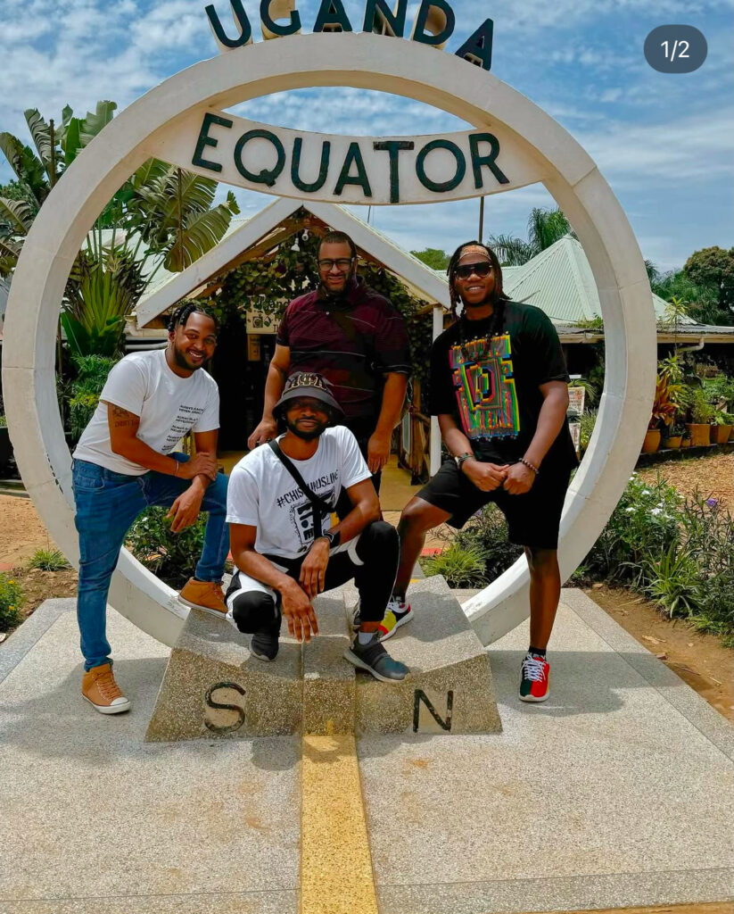 Four men pose at the Uganda Equator landmark, smiling for the camera. Two stand and two crouch in front of the circular sign, with tropical plants and a building in the background.