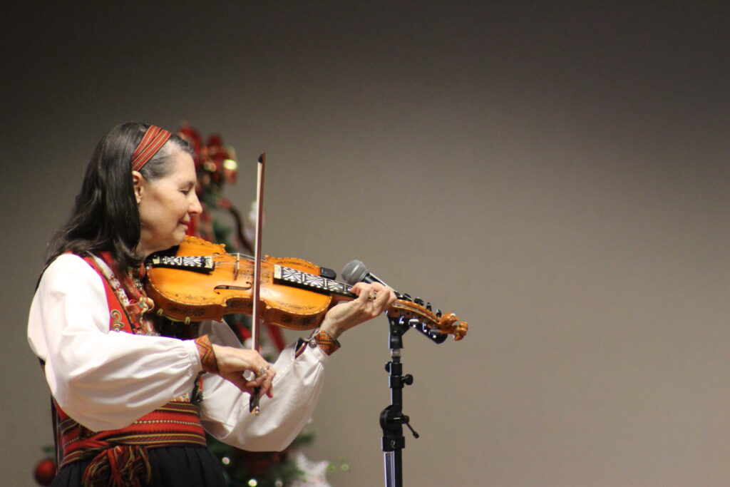 A person wearing a white and red blouse, playing fiddle in front of a dark background.