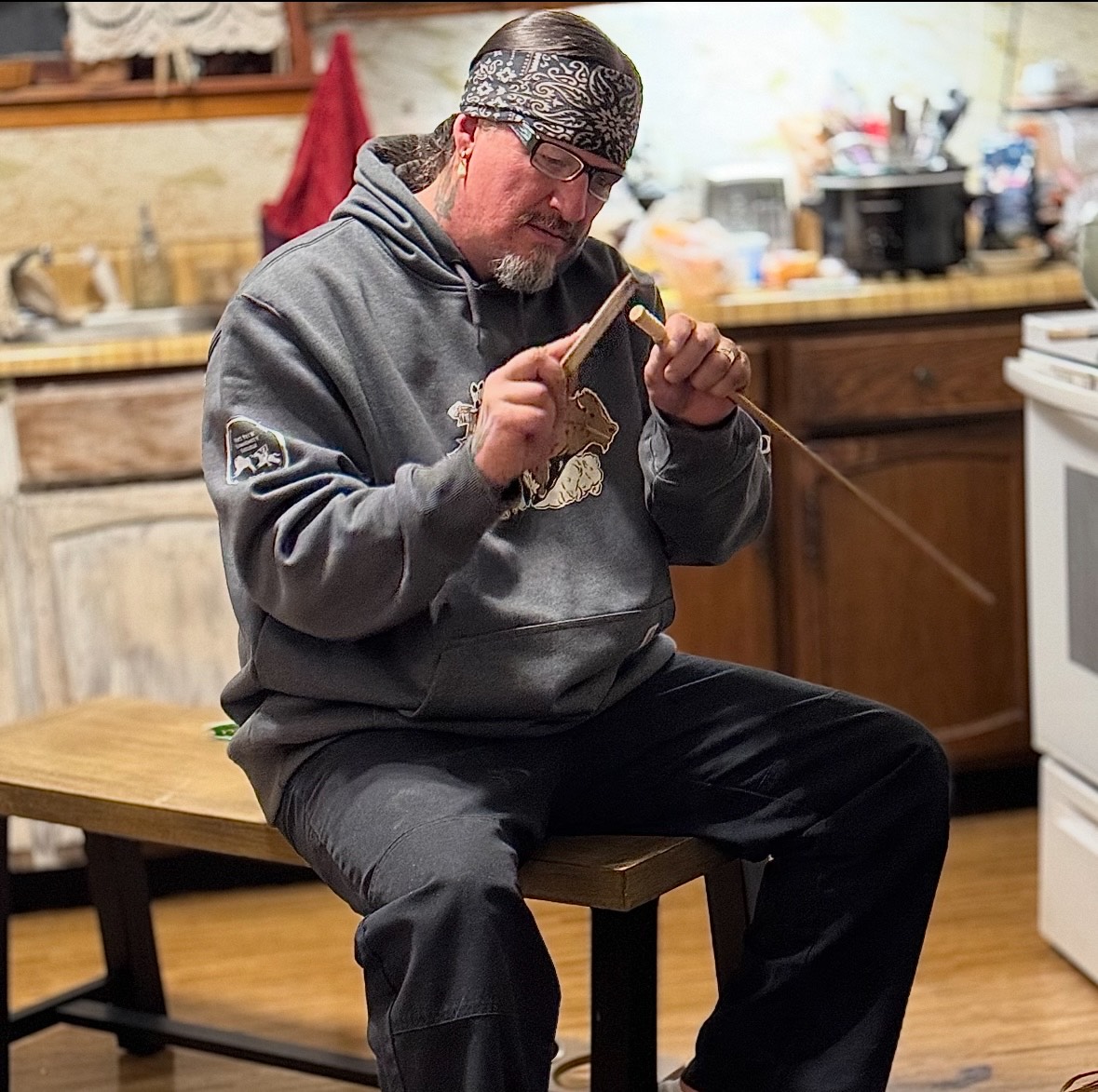 A person sitting on a bench in a kitchen, whittling an arrow.