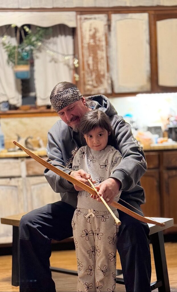 An older person in a kitchen showing a young child how to hold a handcrafted bow and arrow.