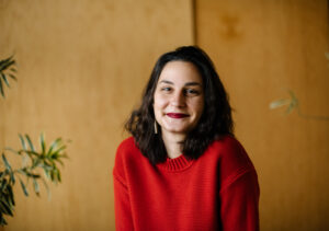 A smiling person of medium light skin tone, with dark brown shoulder length hair, a nose ring, and red lipstick wearing a red sweater.