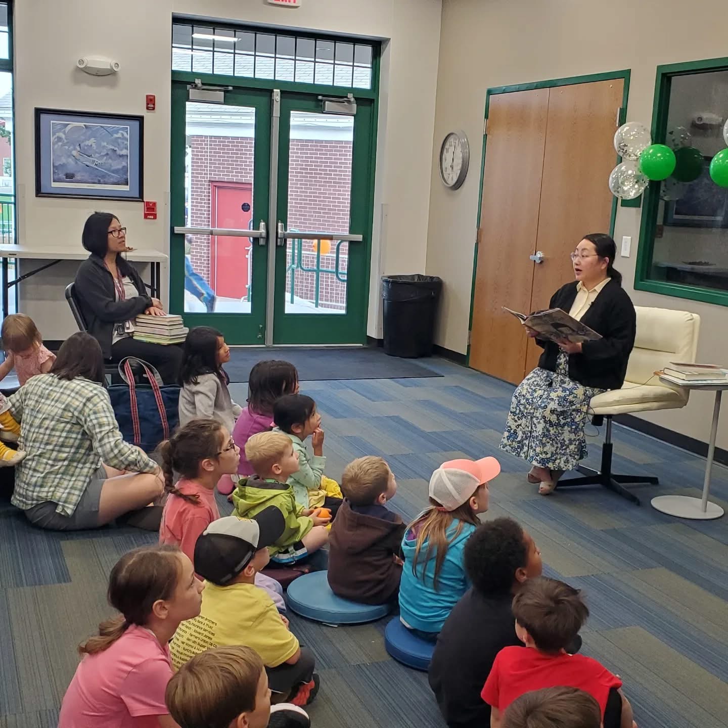 A woman sits in a chair reading a book to a group of children seated on the floor in a library. Another adult sits to the side holding a stack of books