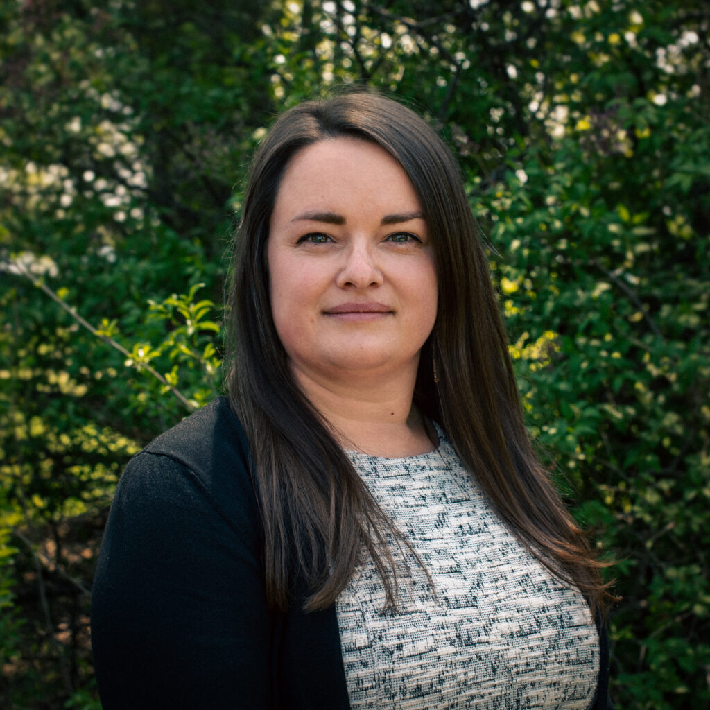 A smiling person of light skin tone with shoulder length brown hair, wearing a light blue top under a black blazer