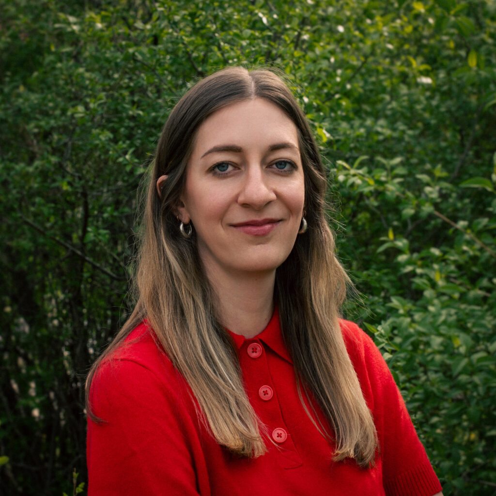 Headshot of a smiling person of light skin tone, with dark blonde long hair, wearing a red shirt.
